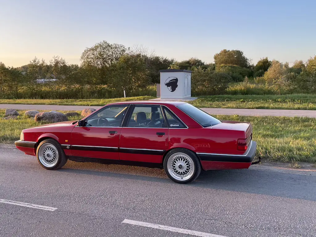 Pildil on päikeseloojangu eel, erkpunane Audi 200 pargituna tee ääres, taustal elektrikilp, millel Edward Von Lõnguse kunstiteos